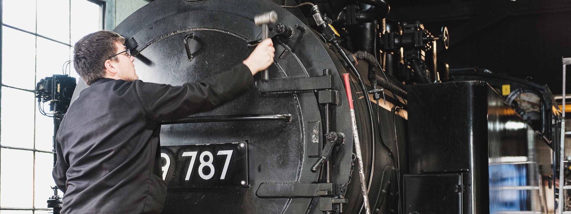 Eine Schwarze Lokomotive von vorne und ein Mann steht davor und prüft etwas mit seinem Hammer.