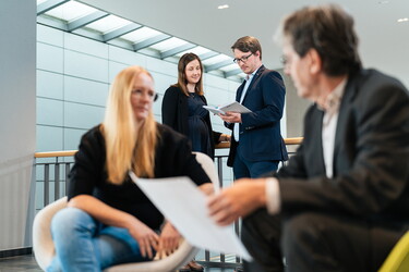 Im Vordergrund sitzen ein Mann und eine Frau im Gespräch. Im Hintergrund stehen ein Mann und eine Frau im Gespräch.
