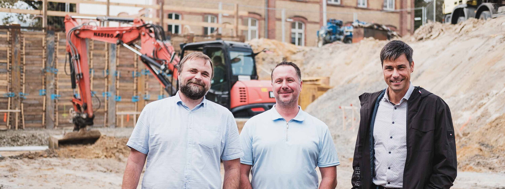 Drei Männer stehen auf einer Baustelle und im Hintergrund steht ein roter Bagger.