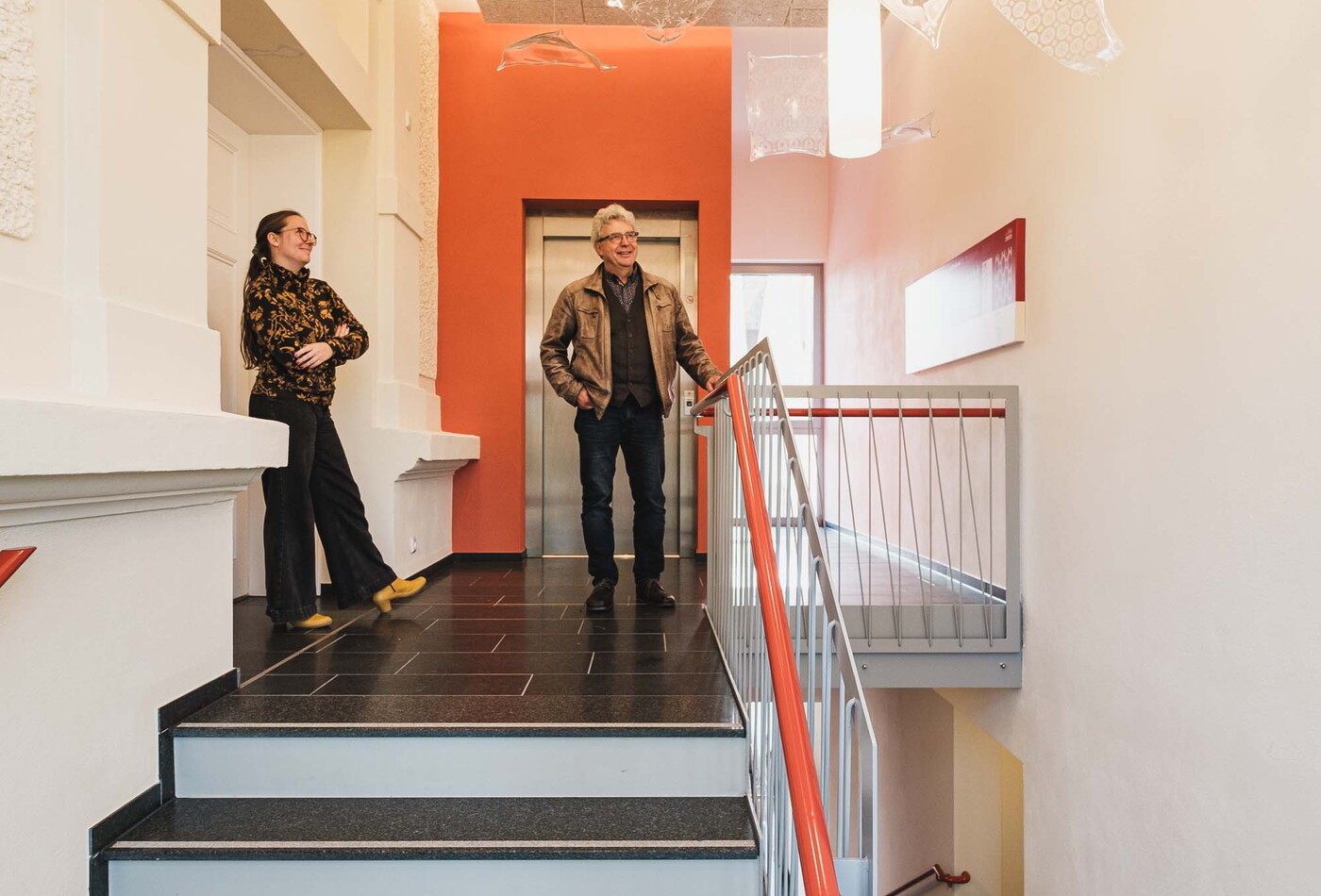 Eine schwarz weiße Treppe und im Flur stehen vor dem Fahrstuhl ein älterer Mann und eine Junge Frau