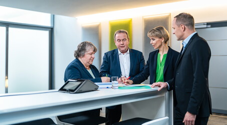 Zwei Männer und zwei Frauen stehen ein einem Tisch und sehen sich Unterlagen an.