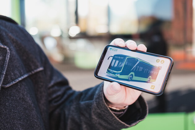 Eine Nahaufnahme wie eine Person ein Handy in den Händen hält und auf dem Handy ist ein grüner Bus abgebildet.
