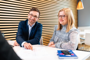 Ein Mann und ein Frau an einem Tisch schauen freundlich zu einer weiteren Person.