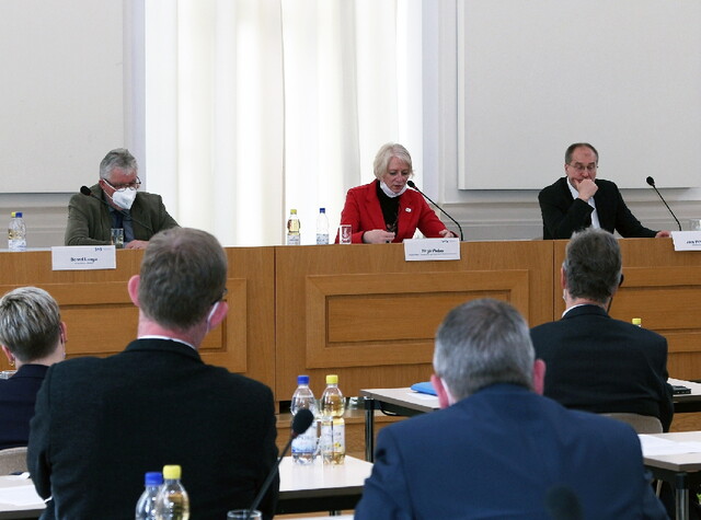 Im Vordergrund sieht man die Rücken der Männer die an ihren Tischen sitzen und im Hintergrund sieht man die Gesichter der Personen am Pult
