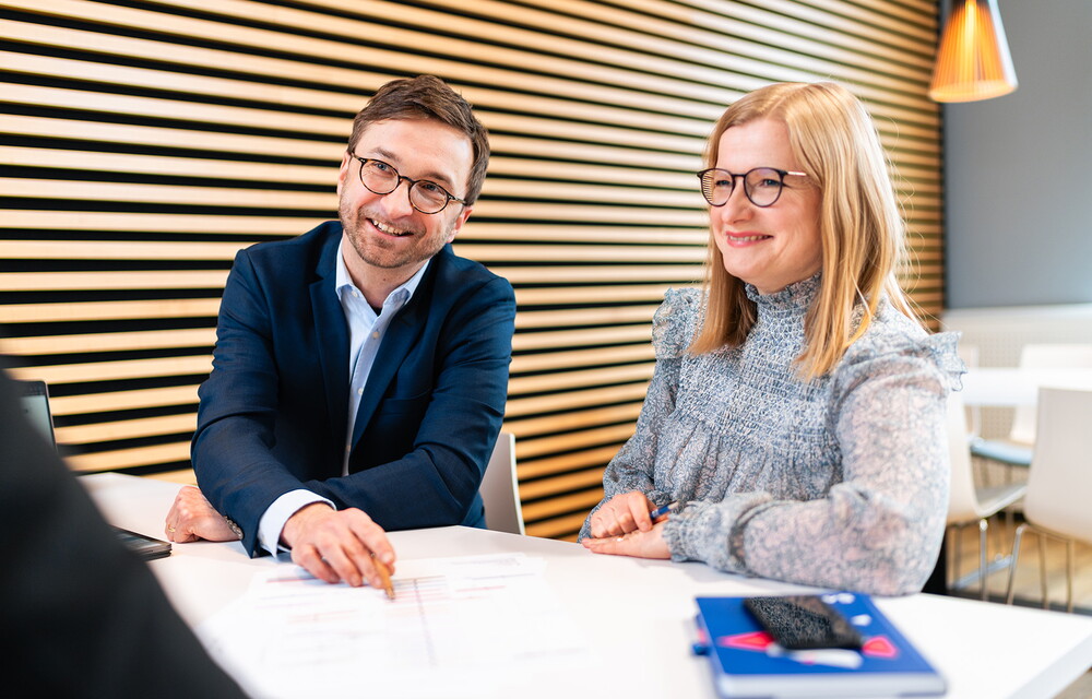 Ein Mann und ein Frau an einem Tisch schauen freundlich zu einer weiteren Person.