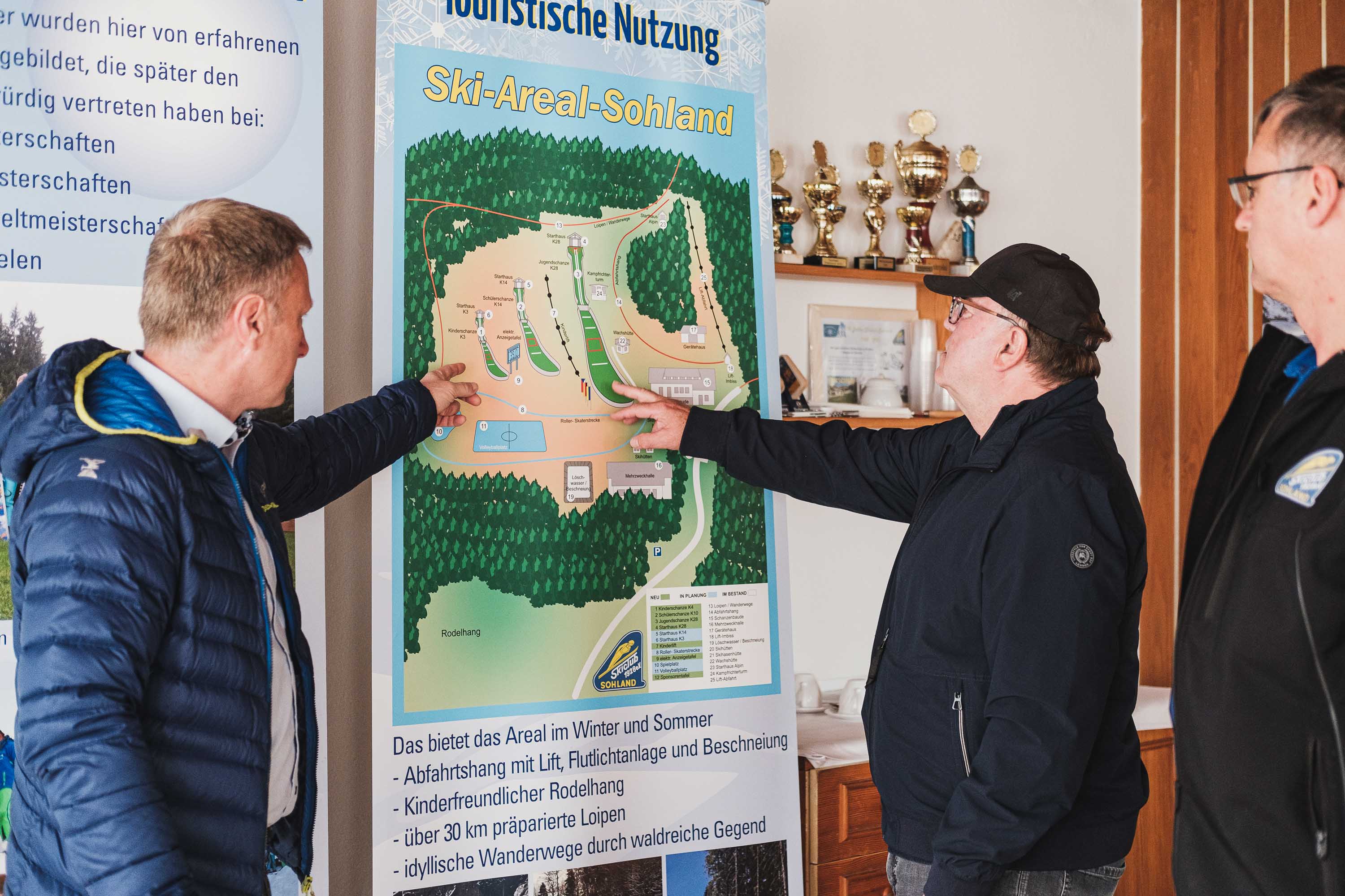 Zwei Männer stehen vor einen Plakat mit der Aufschrift Ski Areal Soland und zweigen auf das Gebiet. Der dritte Mann steht davor und schaut drauf.